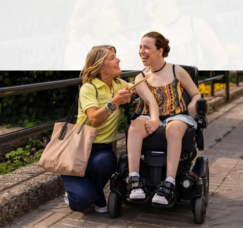 NDIS support worker kneeling beside a joyful young woman in a motorised wheelchair during a community outing in Australia