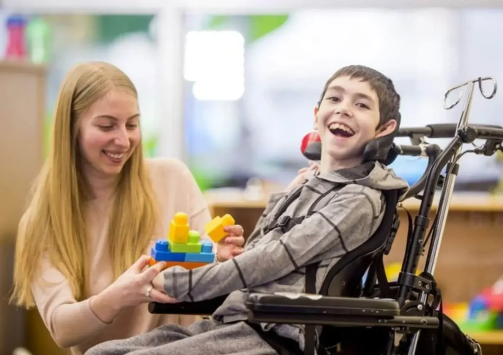 Female NDIS support worker and a smiling child in a wheelchair playing with building blocks during a disability therapy session in Australia