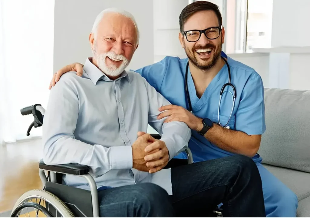 Friendly NDIS support worker in scrubs sitting beside a happy elderly man in a wheelchair, providing aged care and disability support in Australia